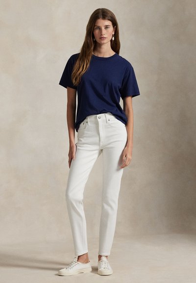 Jonge vrouw draagt een marineblauw T-shirt, witte high-waisted jeans, witte sneakers en pareloorbellen, staand tegen een effen achtergrond.