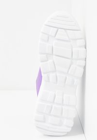 Semelle épaisse blanche à crampons d'une chaussure violette reposant contre un coin de mur et de sol blancs.