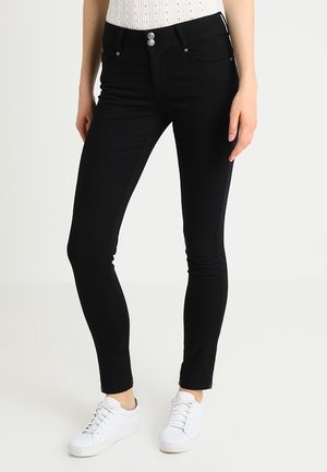 Kvinde iført slanke sorte jeans med dobbelt knaplukning og hvide sneakers, stående mod en ensartet lys baggrund.