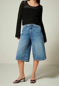 Haut à manches longues noir en maille avec un motif texturé, associé à un pantalon culotte en denim bleu clair. Des sandales à talons noires complètent la tenue.