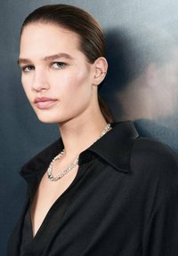 Čierna košeľa s golierom, lesklý strieborný náhrdelník s priehľadnými kameňmi, hladká pokožka, rovné vlasy, neutrálny pozadie. Zameranie na tvár a doplnok.