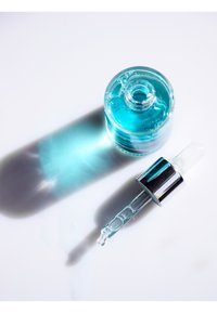 Klar glasflaska med turkosa vätska, silverfärgad droppkappa och avlång dropper. Skugga kastad på vit bakgrund.