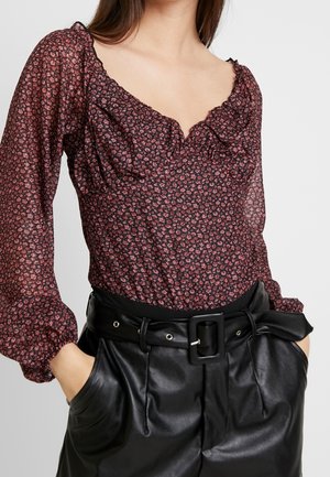 Vrouw die een doorschijnend, lang mouwen bloemenblouse draagt met gerimpelde halslijn en zwarte nepleren broek met een brede riem en grote gesp.