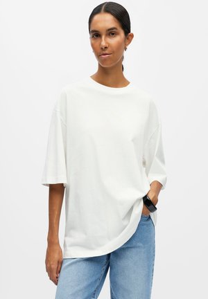 Kvinde med mørkt hår i en lav knold iført en løs hvid kortærmet skjorte, blå jeans og et sort armbånd med den ene hånd i lommen.