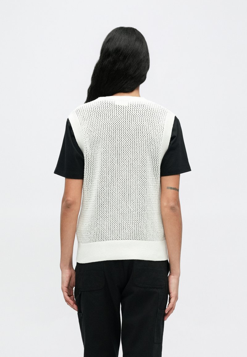 Top bianco senza maniche in maglia con un motivo a trama aperta e testurizzata, indossato sopra una maglietta nera a maniche corte. Pantaloni neri visibili in vita.
