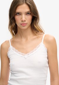 Top de tirantes blanco con costura acanalada y borde de encaje alrededor del escote y las tirantes. Tejido suave, diseño ajustado y delicados acentos de encaje.