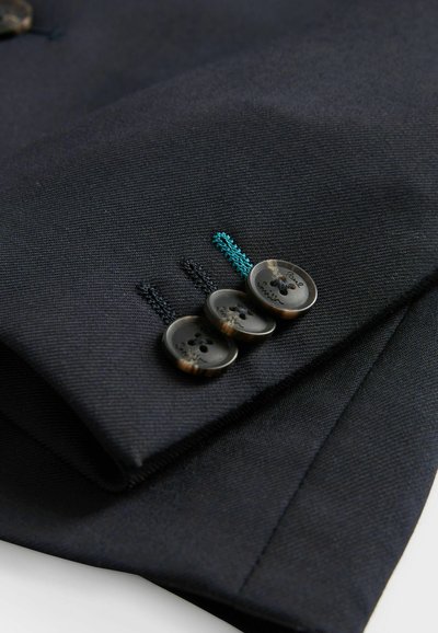 Marinblå tygmanschett med tre mörka knappar, prydd med blågröna och svarta dekorativa stygn. Slät textur, skräddarsydd form och strukturerad finish.