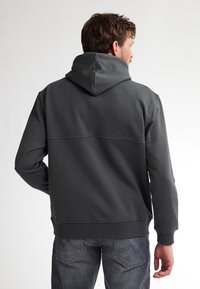 Felpa grigia scura con un taglio rilassato, caratterizzata da un grande cappuccio, polsini a costine e un dettaglio di cucitura sulla parte superiore della schiena. Indossata con jeans scuri.