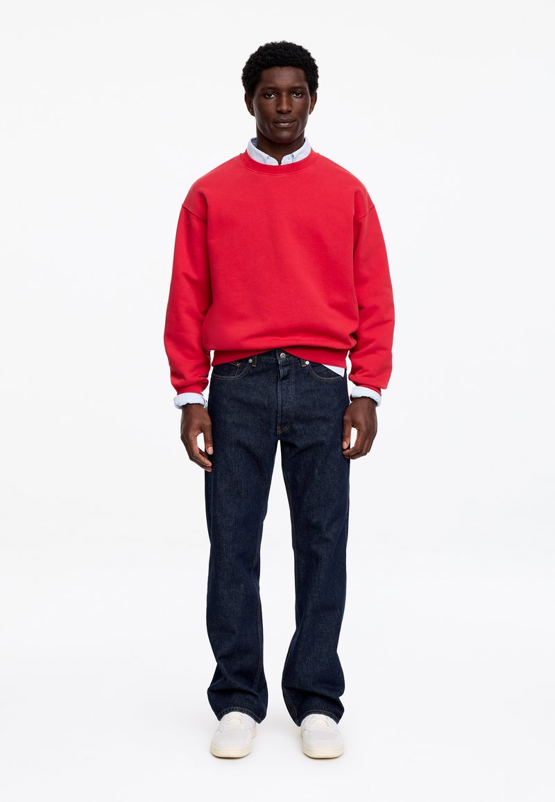 Un homme est debout de face, portant un sweat-shirt rouge par-dessus une chemise blanche à col, un jean bleu foncé et des baskets blanches, sur fond blanc.