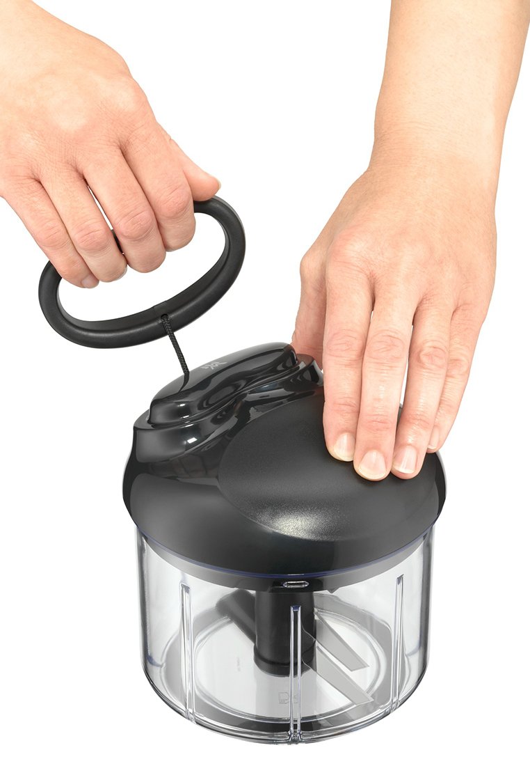 Schwarzer Küchenhacker mit transparentem Behälter. Hat einen runden Deckel, einen ergonomischen Griff und eine sichtbare Klinge im Inneren. Hände greifen den Deckel.