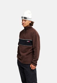 Homme portant un pull polaire marron avec une bande noire, un pantalon noir, un bonnet blanc et des lunettes de soleil sur le bonnet devant un fond uni.