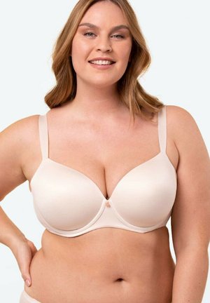 Uśmiechnięta kobieta o włosach do ramion, ubrana w jasnobeżowy biustonosz, stojąca z jedną ręką na biodrze na jednolitym tle.