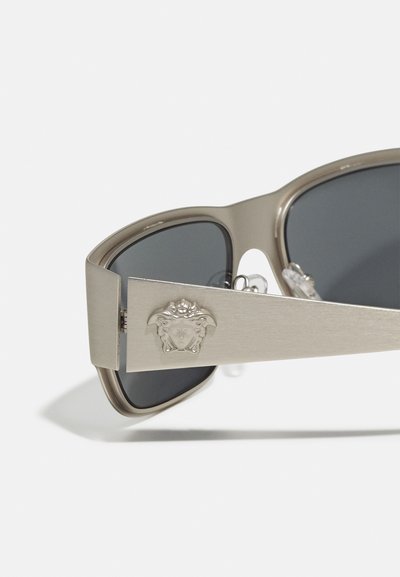 Ochelari de soare cu un cadru metalic argintiu elegant, lentile întunecate și un emblematic cap de leu proeminent pe templu, prezentând un design modern cu decupaje.