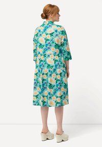 Abito floreale in tessuto turchese con maniche corte, design a strati, con grandi fiori bianchi e gialli, indossato con zoccoli bianchi.