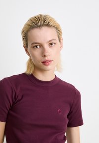 T-shirt a righe bordeaux con collo rotondo e maniche corte, caratterizzato da un piccolo logo sul lato sinistro del petto. Texture liscia.