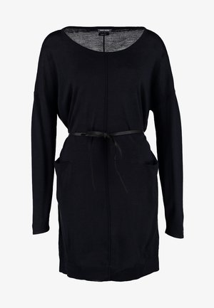 Femme portant une robe noire longue à manches longues ceinturée avec des poches, portant un petit sac noir en bandoulière et des bottines noires à bout ouvert.