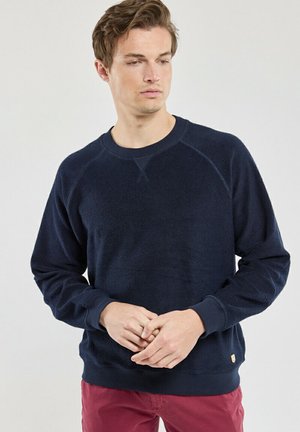 Jeune homme aux cheveux courts et bruns portant un pull bleu marine et un pantalon rouge, regardant vers le bas avec les mains jointes devant lui.