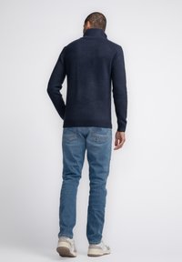 Granatowy sweter dziewiarski z wysokim kołnierzem, prążkowanymi mankietami i dołem. Noszony z jasnoniebieskimi dżinsami i casualowymi sneakersami. Prosty, teksturowany wykończenie.