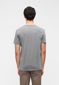 Άνδρας με σκούρα μαλλιά που φοράει απλό γκρι κοντομάνικο μπλουζάκι και μπεζ παντελόνι, στέκεται με την πλάτη προς το μέρος μας μπροστά σε ουδέτερο φόντο.