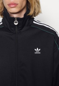 Musta vetoketjullinen takki, jossa korkea kaulus, valkoinen Adidas-logo ja kolme valkoista raitaa olkapäillä sekä turkoosit tikkauskoristeet.