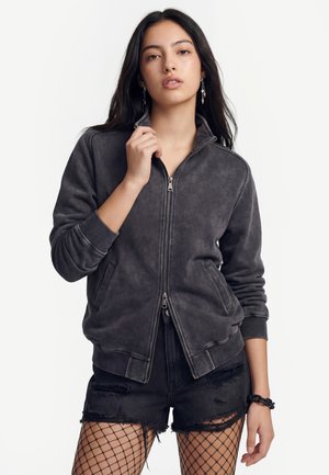 Fiatal nő hosszú sötét hajjal, kopott fekete cipzáras dzsekit, szakadt fekete rövidnadrágot és hálós harisnyát visel, a dzseki gallérját fogja.