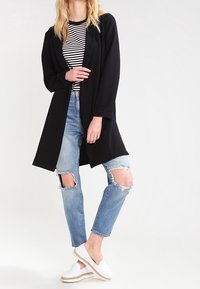 Kobieta w czarnej kurtce rozpinanej z przodu, w czarno-białą pasiastą koszulę, w jasnoniebieskie podarte dżinsy i białe mokasyny, stojąca na jednej nodze.