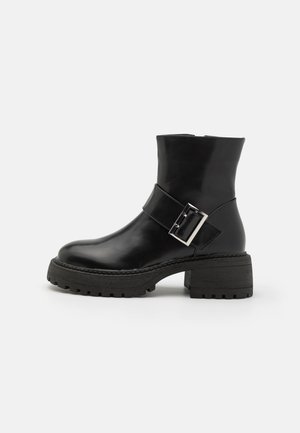 Bottines en cuir noir avec un talon bloc, ornées d'une sangle décorative et d'un détail de boucle, avec une semelle texturée et une finition lisse.