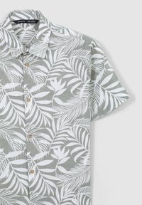Camicia a maniche corte con bottoni, realizzata in tessuto leggero, con uno sfondo verde e motivi di foglie tropicali bianche e bottoni in legno.