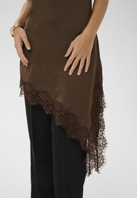 Haut asymétrique marron avec bordure en dentelle et détails à franges ; matière lisse ; associé à un pantalon noir ; mains posées sur l'ourlet.