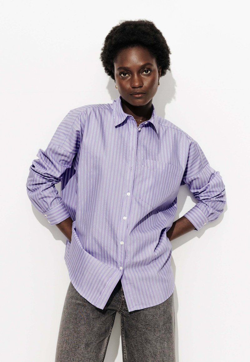 Chemise rayée à boutons de couleur violet clair avec des rayures blanches verticales, dotée d'un col et de manches longues retroussées. Poche sur la poitrine.