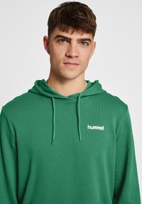 Grön hoodie gjord av mjukt material, med dragsko i huvan och en vit logotyp "hummel" på bröstet. Slät textur, avslappnad design.
