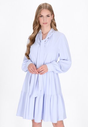 Vaaleansininen mekko, jossa on pitkät hihat, solmittava kaula-aukko ja kerroksellinen röyhelöhelma. Valmistettu sileästä kankaasta, jossa on virtaava siluetti.