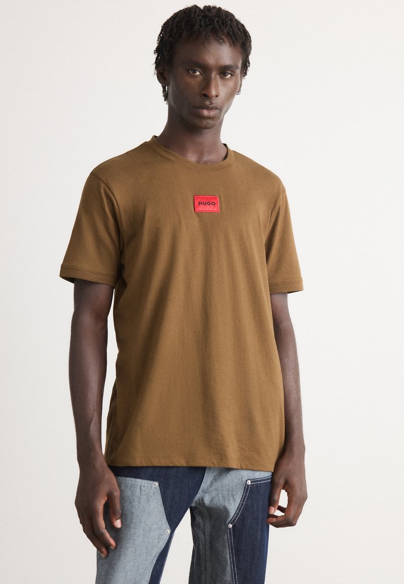 Ung mann med korte dreadlocks iført en brun T-skjorte med rød Hugo-merke og jeans i to farger, stående mot en ensfarget bakgrunn.