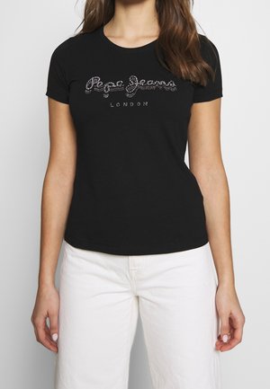 Frau trägt ein eng anliegendes schwarzes T-Shirt mit silbernen Nieten und dem Schriftzug „Pepe Jeans London“, kombiniert mit hoch taillierten weißen Hosen.