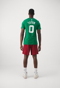 Man met zijn rug naar de camera, gekleed in een groen "Tatum 0" shirt, rode shorts, witte Nike sokken en witte sneakers, tegen een effen lichte achtergrond.