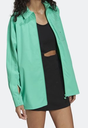 Femme portant une veste surdimensionnée turquoise vif sur un haut court noir et une jupe noire, debout devant un fond blanc.