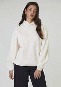 Jonge vrouw met lang donker haar, die gouden ringoorbellen, een off-white hoodie en zwarte broeken draagt, staand tegen een effen achtergrond.