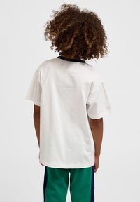 Göndör hajú gyerek hátulnézetben, laza fehér pólót és zöld nadrágot visel tengerészkék oldalsávokkal, egyszerű háttér előtt.