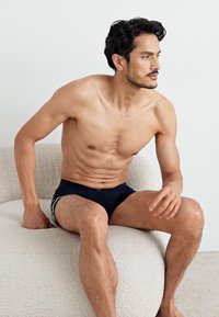 Zwarte zwemboxers met witte accenten gedragen door een zittende man met kort, donker haar. Gladde huid en een lichte achtergrond; ronde, zachte stoel.