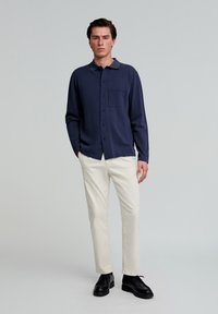 Cămașă albastră cu nasturi, cu guler și buzunar frontal, asociată cu pantaloni de culoare crem și pantofi negri. Design simplu și aerodinamic.