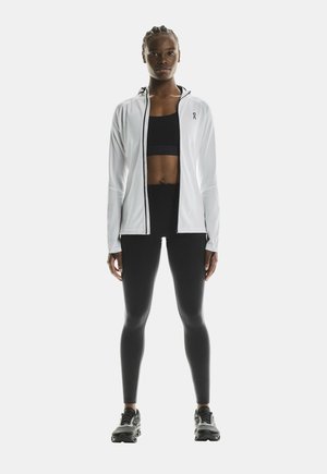 Athletic outfit featuring a white zip-up jacket with black accents, black sports bra, and black leggings, complemented by black athletic shoes.