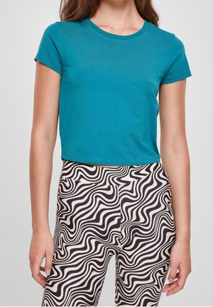 Kvinde iført teal kortærmet crop top og sort-hvide bølgede mønstrede bukser med høj talje, stående mod en ensfarvet baggrund.