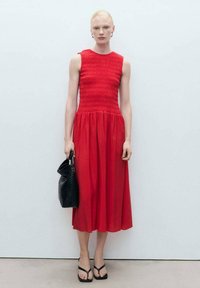 Robe rouge sans manches avec un corsage smocké, jupe fluide, portée avec des tongs noires et un sac à main noir texturé. Tissu lisse.