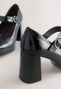 Black patent leather Mary Jane shoes with perforated detailing, round silver buckles, and chunky high block heels on a light background.
