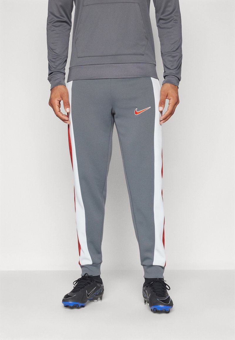 Homme portant un pantalon de survêtement gris avec des bandes latérales blanches et rouges, un sweat à capuche gris et des crampons noirs avec des semelles bleues.