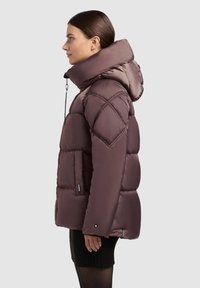 Kvinde i profil iført en brun polstret jakke med høj krave og hætte, parret med en sort nederdel og strømpebukser mod en neutral baggrund.