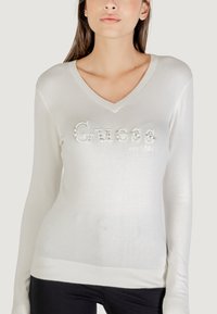 Witte V-hals trui met lange mouwen, voorzien van een verhoogd 'Guess' logo dat versierd is met strass en geribbelde boorden en zoom.