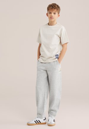 Jongen staat met handen in de zakken, draagt een beige t-shirt, grijze joggingbroek en witte sneakers met zwarte strepen tegen een effen achtergrond.