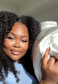 Model with curly black hair holds a white satin pillow. She wears a light blue top. The lighting is bright, highlighting her features.