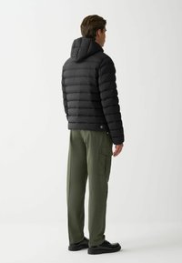 Melna pūkainā jaka ar horizontālu quiltētu dizainu un kapuci, kopā ar olīvzaļām kravas biksēm ar sānu kabatām un pogu detaļu.